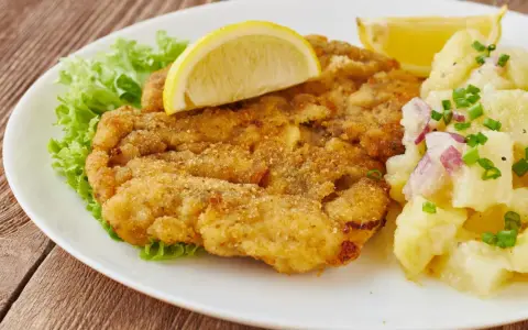 Ein knuspriges Wiener Schnitzel mit Zitrone, Salat und Kartoffelsalat auf einem weißen Teller.
