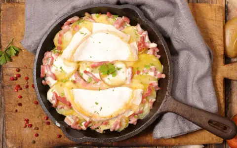 Herzhaftes Tartiflette-Rezept in Gusseisenpfanne: Kartoffeln, Speck und geschmolzener Käse, garniert mit Petersilie.