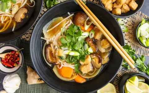 Herzhaftes Pho Chay mit Udon-Nudeln, Shiitake-Pilzen, Tofu und frischem Koriander. Daneben kleine Schälchen mit Chili, Lauchzwiebeln und Limettenspalten.