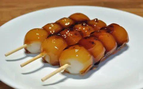 Drei Spieße mit süßen, glasierten japanischen Desserts, die auf einem weißen Teller liegen.