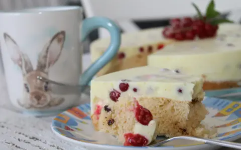 Ein Stück Johannisbeertorte mit Joghurt, dekoriert mit frischen roten Johannisbeeren. Daneben eine Tasse mit Hasenmotiv.