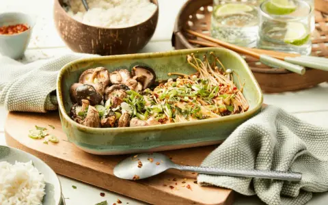 Schüssel mit gebratenen asiatischen Pilzen, garniert mit Frühlingszwiebeln und Koriander, daneben Reis und Chilis.