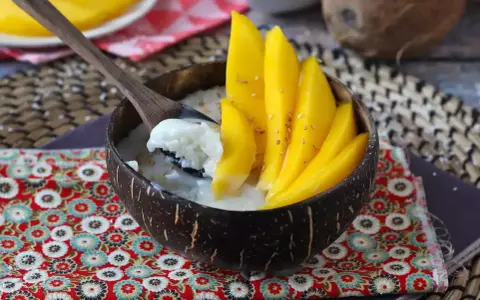 Ein Schälchen mit Mango Sticky Rice, garniert mit frischen Mangoscheiben und Sesam. Ein Löffel hebt cremigen Klebreis mit Kokosmilch hervor.