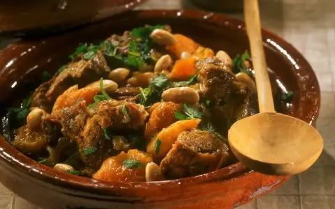 Marokkanisches Lamm mit Aprikosen, Mandeln und frischer Minze in einem traditionellen Tajine-Topf, serviert mit einem Holzlöffel.