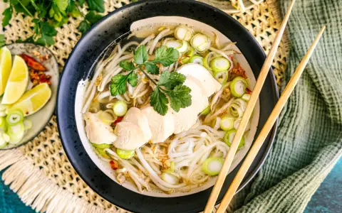 Eine dampfende Schüssel Pho Ga mit zarten Hühnerbrustscheiben, Reisnudeln, Sojasprossen und Frühlingszwiebeln. Serviert mit Limettenspalten und Chili.