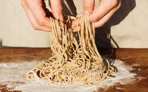 Hände halten frisch gemachte Ramen-Nudeln über einem Holztisch mit Mehl. So einfach kann ramen selber machen sein!