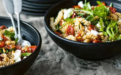 Zwei Schalen mit einem gesunden Nudelsalat aus Spiralnudeln, getrockneten Tomaten, Pinienkernen, Rucola und Feta.