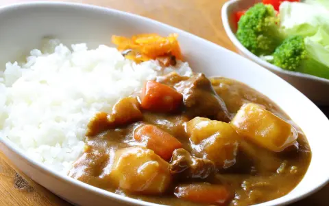 Ein Teller mit dampfendem Reis und herzhaftem Curry, das japanische Nationalgericht, mit zarten Fleischstücken, Kartoffeln und Karotten. Daneben ein kleiner Salat.