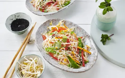 Frischer Glasnudelsalat mit knackigem Gemüse, garniert mit Sesam und Minze. Daneben Schüsselchen mit Sprossen und schwarzem Sesam.