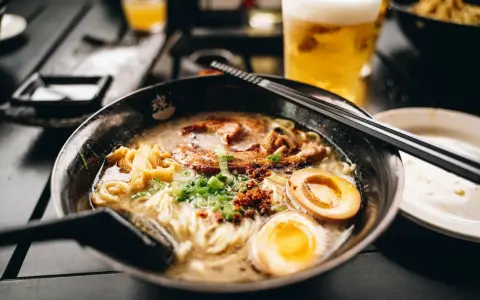 Eine dampfende Schüssel mit den besten Ramen-Bowls, gefüllt mit Nudeln, Schweinefleisch, Ei und Frühlingszwiebeln. Daneben ein Glas Bier.