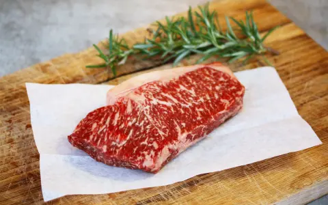 Ein rohes Stück Wagyu-Fleisch mit feiner Marmorierung liegt auf Backpapier auf einem Holzbrett. Ein Rosmarinzweig liegt daneben.