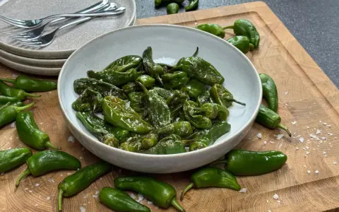 Schüssel mit gebratenen Pimientos de Padrón, bestreut mit grobem Salz. Einige rohe Paprika liegen daneben auf einem Holzbrett.
