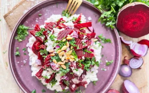 Roter Bete Salat mit cremigem Käse, Pinienkernen und frischer Petersilie auf einem lila Teller.