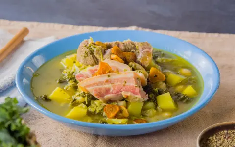 Herzhafter spanischer Eintopf mit Fleisch, Kartoffeln, Grünkohl und Karotten in einer blauen Schüssel.