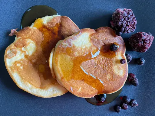 Zwei goldbraune Pancakes ohne Backpulver, beträufelt mit Sirup und garniert mit gefrorenen Beeren auf dunklem Teller.