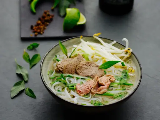 Eine dampfende Schüssel Pho Suppe mit zarten Rindfleischscheiben, Reisnudeln, Sojasprossen und frischen Kräutern. Ein Hauch von Limette und Gewürzen im Hintergrund.