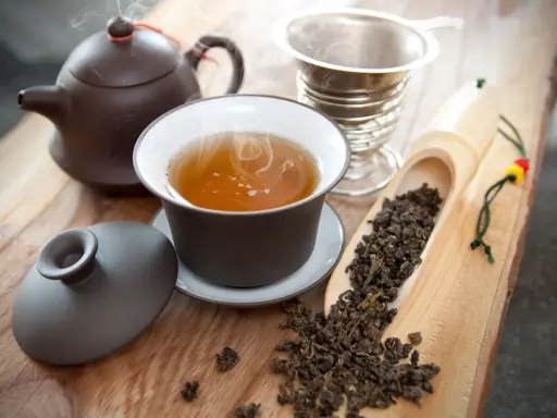 Dampf steigt aus einer Tasse Oolong Tee auf. Daneben liegen lose Teeblätter in einer Holzschaufel. So schmeckt Oolong Tee: mild, blumig, leicht süßlich.