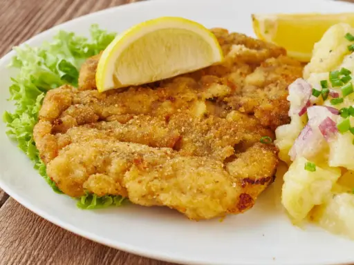 Ein knuspriges Wiener Schnitzel mit Zitrone, Salat und Kartoffelsalat auf einem weißen Teller.