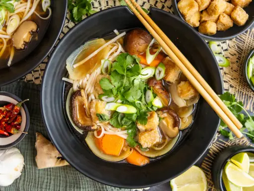 Herzhaftes Pho Chay mit Udon-Nudeln, Shiitake-Pilzen, Tofu und frischem Koriander. Daneben kleine Schälchen mit Chili, Lauchzwiebeln und Limettenspalten.