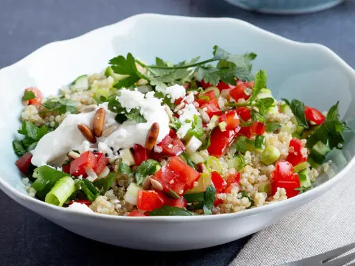 Frischer Quinoa-Salat mit Feta, Tomaten, Gurken und Kräutern, garniert mit Joghurt und Pinienkernen.
