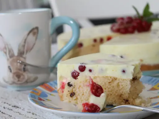 Ein Stück Johannisbeertorte mit Joghurt, dekoriert mit frischen roten Johannisbeeren. Daneben eine Tasse mit Hasenmotiv.