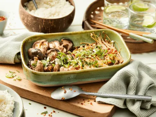 Schüssel mit gebratenen asiatischen Pilzen, garniert mit Frühlingszwiebeln und Koriander, daneben Reis und Chilis.