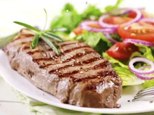 Ein saftiges Steak mit Grillstreifen, garniert mit Rosmarin, neben einem frischen Salat aus knackigem Grün, Tomaten und roten Zwiebelringen. Ein perfekter Steak Salat.