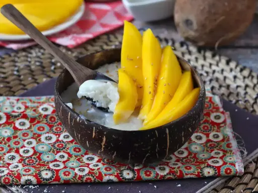 Ein Schälchen mit Mango Sticky Rice, garniert mit frischen Mangoscheiben und Sesam. Ein Löffel hebt cremigen Klebreis mit Kokosmilch hervor.