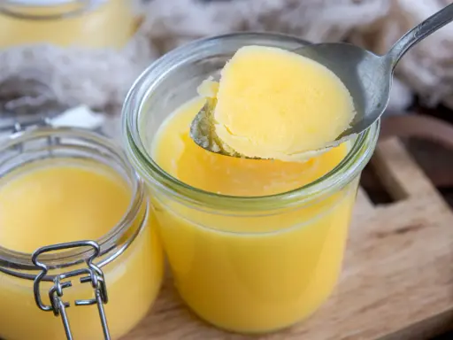Ein Löffel Butterschmalz wird aus einem Glas gehoben. Dieses goldgelbe Fett ist ideal zum Braten, da sein hoher Butterschmalz rauchpunkt Verbrennungen verhindert.