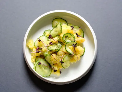 Kartoffelsalat mit frischer Gurke, garniert mit lila Sprossen und Dill. Ein leichter und erfrischender Genuss.