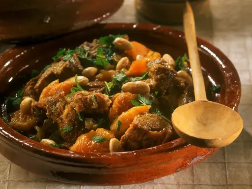 Marokkanisches Lamm mit Aprikosen, Mandeln und frischer Minze in einem traditionellen Tajine-Topf, serviert mit einem Holzlöffel.