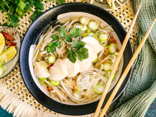 Eine dampfende Schüssel Pho Ga mit zarten Hühnerbrustscheiben, Reisnudeln, Sojasprossen und Frühlingszwiebeln. Serviert mit Limettenspalten und Chili.