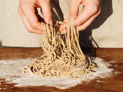 Hände halten frisch gemachte Ramen-Nudeln über einem Holztisch mit Mehl. So einfach kann ramen selber machen sein!