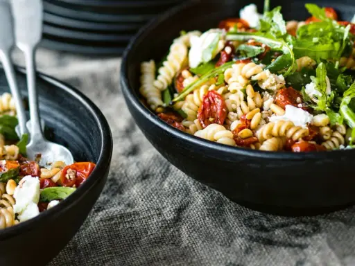 Zwei Schalen mit einem gesunden Nudelsalat aus Spiralnudeln, getrockneten Tomaten, Pinienkernen, Rucola und Feta.