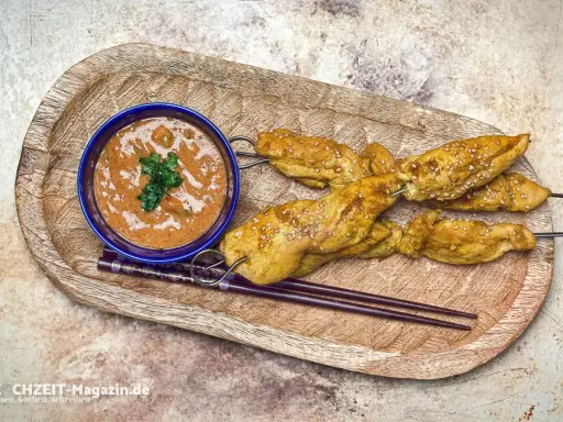 Saftige Satay Chicken Spieße mit Sesam, serviert mit Erdnusssauce und Stäbchen auf einem Holztablett.