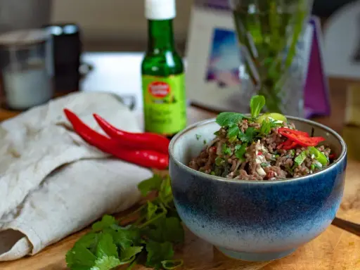 Würziges Laab Moo, ein thailändischer Hackfleischsalat, garniert mit frischer Minze und Chili. Daneben liegen rote Chilis und Koriander.