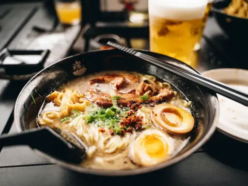 Eine dampfende Schüssel mit den besten Ramen-Bowls, gefüllt mit Nudeln, Schweinefleisch, Ei und Frühlingszwiebeln. Daneben ein Glas Bier.