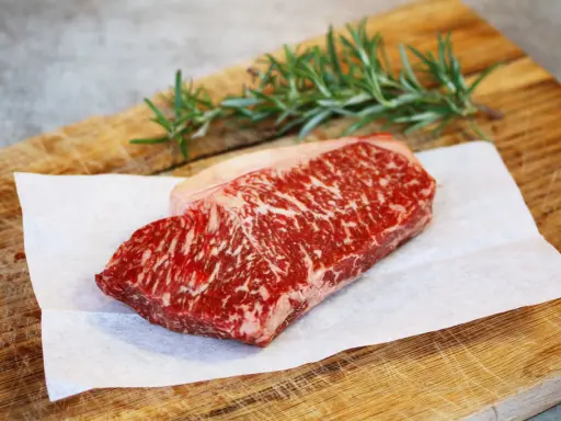 Ein rohes Stück Wagyu-Fleisch mit feiner Marmorierung liegt auf Backpapier auf einem Holzbrett. Ein Rosmarinzweig liegt daneben.