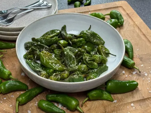 Schüssel mit gebratenen Pimientos de Padrón, bestreut mit grobem Salz. Einige rohe Paprika liegen daneben auf einem Holzbrett.