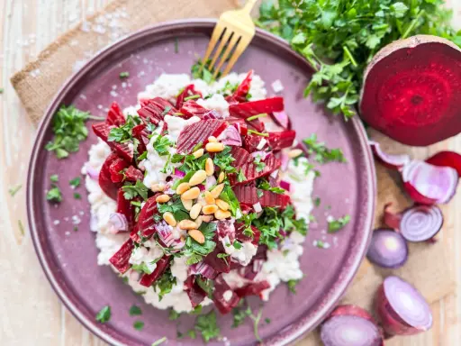 Roter Bete Salat mit cremigem Käse, Pinienkernen und frischer Petersilie auf einem lila Teller.