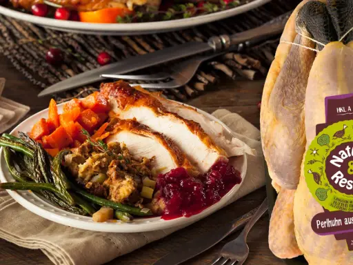 Ein festlich gedeckter Tisch mit einem Teller voller gebratenem Perlhuhn, dazu grüne Bohnen, Karotten und Cranberry-Sauce. Das unzubereitete Perlhuhn daneben zeigt das Label "Nature & Respect".