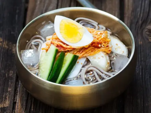 Erfrischendes Mul Naengmyeon mit Buchweizennudeln, Kimchi, Gurke, Ei und Eiswürfeln in einer bronzenen Schüssel auf Holzuntergrund.