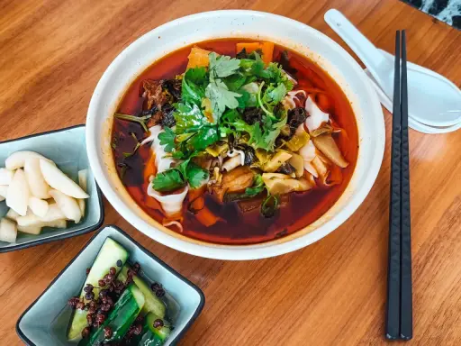 Eine Schüssel mit aromatischer taiwanischer Rindernudelsuppe, garniert mit frischem Koriander, daneben eingelegte Gurken und Rettich.