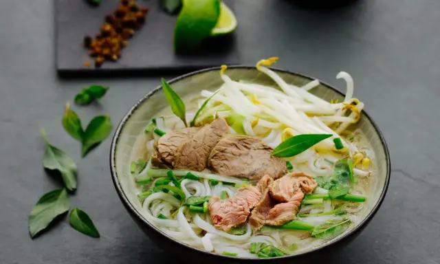 Eine dampfende Schüssel Pho Suppe mit zarten Rindfleischscheiben, Reisnudeln, Sojasprossen und frischen Kräutern. Ein Hauch von Limette und Gewürzen im Hintergrund.
