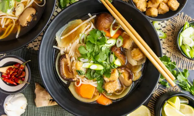 Herzhaftes Pho Chay mit Udon-Nudeln, Shiitake-Pilzen, Tofu und frischem Koriander. Daneben kleine Schälchen mit Chili, Lauchzwiebeln und Limettenspalten.