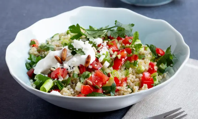 Frischer Quinoa-Salat mit Feta, Tomaten, Gurken und Kräutern, garniert mit Joghurt und Pinienkernen.