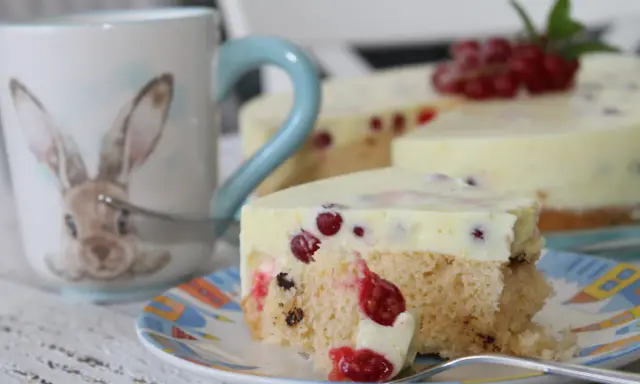 Ein Stück Johannisbeertorte mit Joghurt, dekoriert mit frischen roten Johannisbeeren. Daneben eine Tasse mit Hasenmotiv.