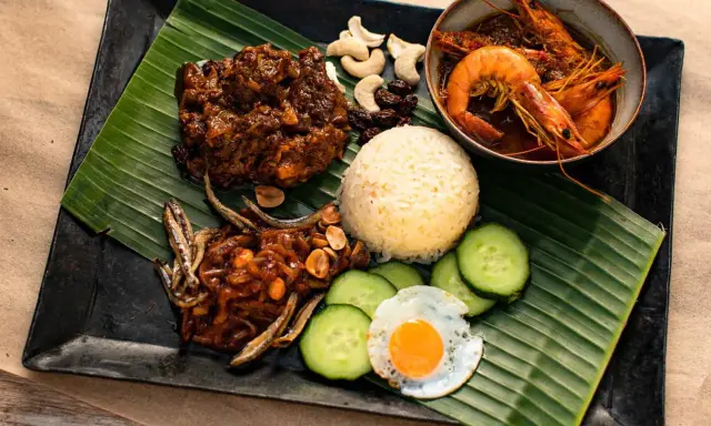 Nasi Lemak: Reis, scharfe Garnelen, frittierte Sardellen, Spiegelei, Gurkenscheiben, Cashewkerne und Rosinen auf Bananenblatt.