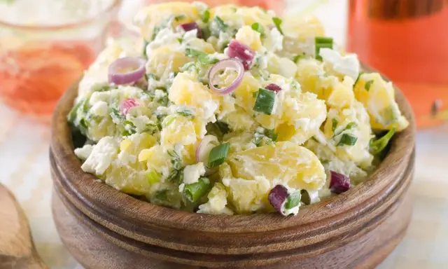 Herzhafter Kartoffelsalat mit Meerrettich, verfeinert mit Frühlingszwiebeln und roten Zwiebelringen, serviert in einer Holzschale.