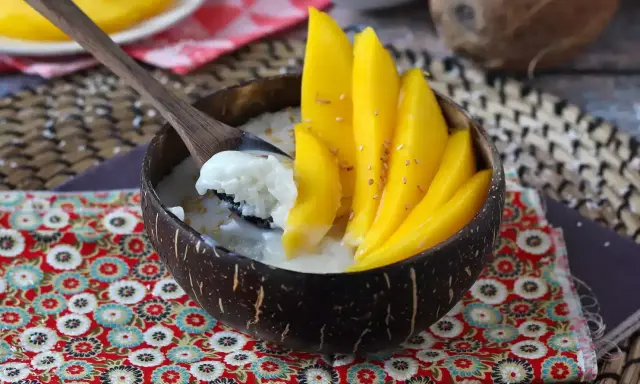 Ein Schälchen mit Mango Sticky Rice, garniert mit frischen Mangoscheiben und Sesam. Ein Löffel hebt cremigen Klebreis mit Kokosmilch hervor.